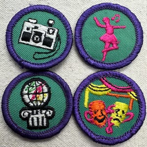May include: Four circular patches with green centers and purple borders. The patches feature embroidered designs: a camera, a dancer, a globe on a column, and two theater masks. The designs are in white, pink, orange, and yellow.