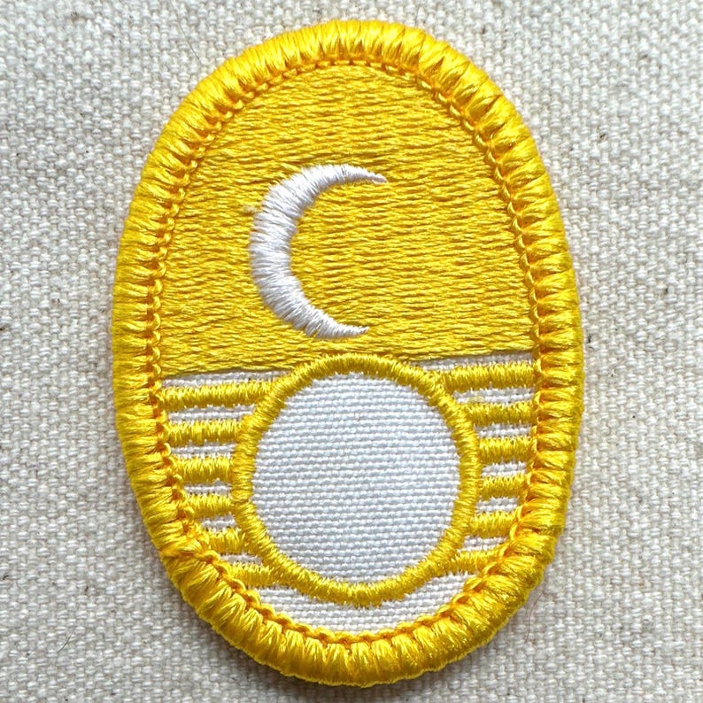 Puede incluir: Parche bordado amarillo con un dise&ntilde;o de luna creciente blanca y c&iacute;rculo blanco. El parche tiene un borde cosido detallado y una apariencia texturizada, lo que sugiere un art&iacute;culo artesanal.