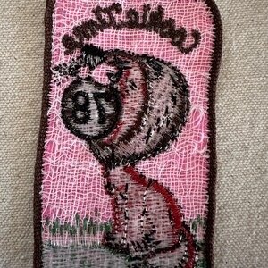 Puede incluir: Parche bordado con un personaje de dibujos animados y el texto "Smutty Babe". El parche tiene un fondo rosa y un borde marr&oacute;n. El personaje es en tonos marrones, rojos y negros, con el n&uacute;mero "87" en su cuerpo.