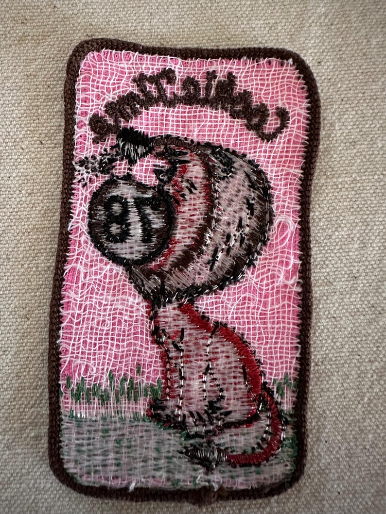 Puede incluir: Parche bordado con un personaje de dibujos animados y el texto "Smutty Babe". El parche tiene un fondo rosa y un borde marr&oacute;n. El personaje es en tonos marrones, rojos y negros, con el n&uacute;mero "87" en su cuerpo.