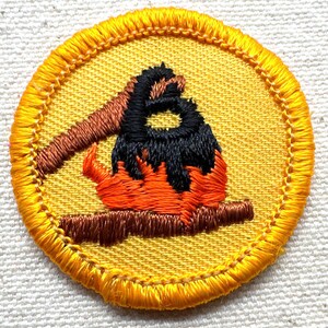 May include: Embroidered patch featuring a campfire scene. The patch has a yellow background with a detailed image of a campfire in orange, black, and brown. The patch is round with a yellow stitched border. Ideal for scouts or outdoor enthusiasts.