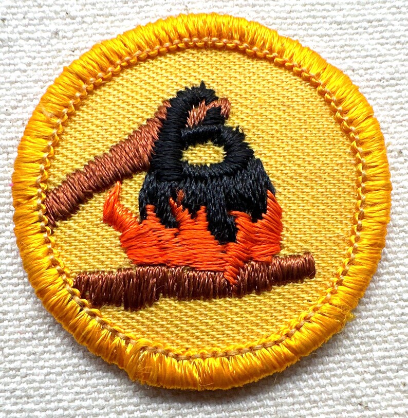 May include: Embroidered patch featuring a campfire scene. The patch has a yellow background with a detailed image of a campfire in orange, black, and brown. The patch is round with a yellow stitched border. Ideal for scouts or outdoor enthusiasts.