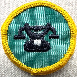 May include: Embroidered patch with a green background and a black and white design in the center. The patch has a yellow border. The design appears to be a stylized representation of a balance scale.