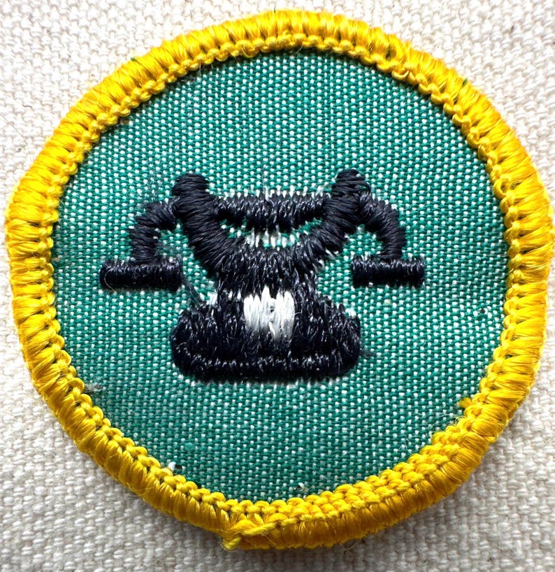 May include: Embroidered patch with a green background and a black and white design in the center. The patch has a yellow border. The design appears to be a stylized representation of a balance scale.