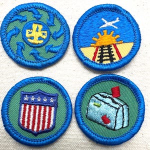 May include: Four embroidered patches with blue borders. The patches feature various designs, including a recycling symbol, a train with a sun and airplane, a shield with stars and stripes, and a building with a red chimney. The patches are on a green background.