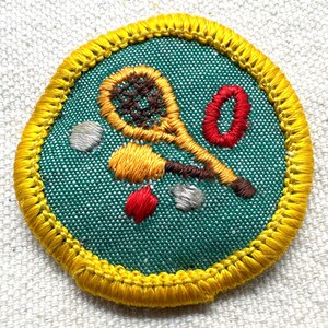 May include: Embroidered patch with a tennis racket, tennis balls, and a red zero on a teal background. The patch is round with a yellow border. The tennis racket is yellow and brown, and the tennis balls are yellow, red, and gray.