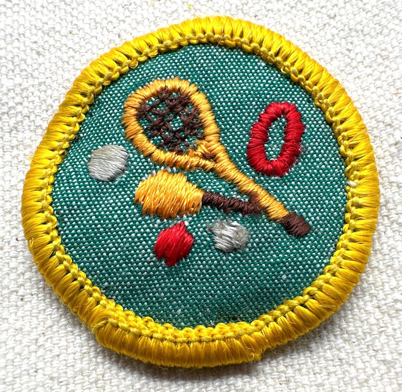May include: Embroidered patch with a tennis racket, tennis balls, and a red zero on a teal background. The patch is round with a yellow border. The tennis racket is yellow and brown, and the tennis balls are yellow, red, and gray.