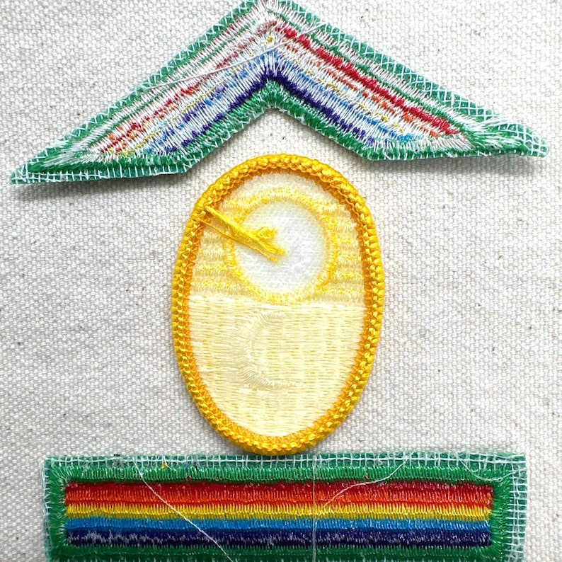 Puede incluir: Tres parches bordados: un gal&oacute;n verde y multicolor, un &oacute;valo amarillo con un dise&ntilde;o de sol y un parche rectangular verde con rayas arco&iacute;ris. Los parches est&aacute;n sobre una tela texturizada blanquecina.