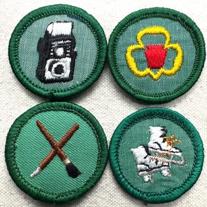 May include: Four round green patches with embroidered designs. One patch features a camera, another a yellow and red emblem, a third crossed paintbrushes, and the last a white and brown design.