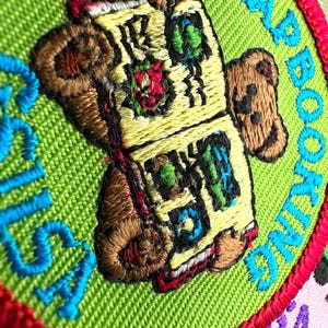 May include: Embroidered patch with the words "SCRAP BOOKING" in turquoise on a green background. The patch features a teddy bear holding a book with colorful details. The patch is bordered with red thread.