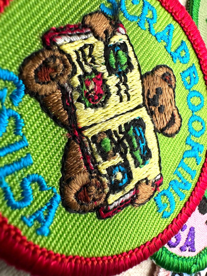 May include: Embroidered patch with the words "SCRAP BOOKING" in turquoise on a green background. The patch features a teddy bear holding a book with colorful details. The patch is bordered with red thread.