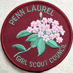May include: A round, embroidered patch with the words "PENN LAUREL GIRL SCOUT COUNCIL" in green. The patch features a cluster of pink flowers with green leaves on a maroon background, and a red border.