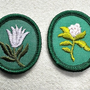 May include: Two embroidered patches with floral designs. One features a white tulip, the other a cluster of small pink flowers. Both are set on a green oval background with a green stitched border. The patches are likely for clothing or crafts.