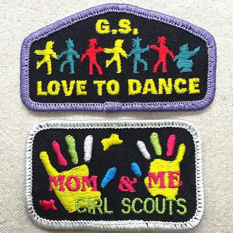 May include: Two embroidered patches. The top patch is black with a purple border and features the text "G.S." and "LOVE TO DANCE" with dancing figures. The bottom patch is black with a white border and reads "MOM & ME GIRL SCOUTS" with handprints.