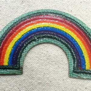 May include: Embroidered rainbow patch with a green border. The rainbow features bands of red, orange, yellow, green, blue, and purple. The patch is on a light-colored fabric background.