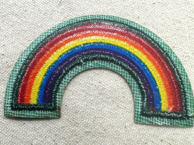 May include: Embroidered rainbow patch with a green border. The rainbow features bands of red, orange, yellow, green, blue, and purple. The patch is on a light-colored fabric background.