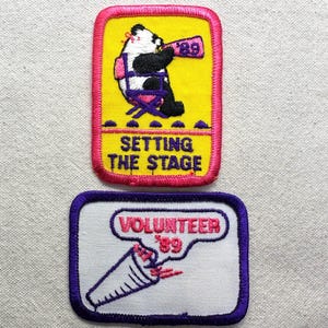 May include: Two rectangular embroidered patches. The top patch has a yellow background with a pink border, featuring a panda sitting on a purple chair holding a megaphone with '89' and the text 'SETTING THE STAGE'. The bottom patch is white with a purple border, featuring a megaphone graphic and the text 'VOLUNTEER '89'.