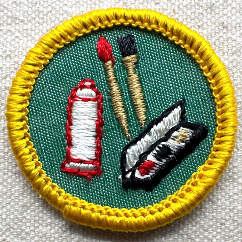 May include: Embroidered patch with a green center and a yellow border. The patch features a red and white paint tube, two paint brushes, and a palette with red, black, and white paint. The patch is likely for an art-related activity.