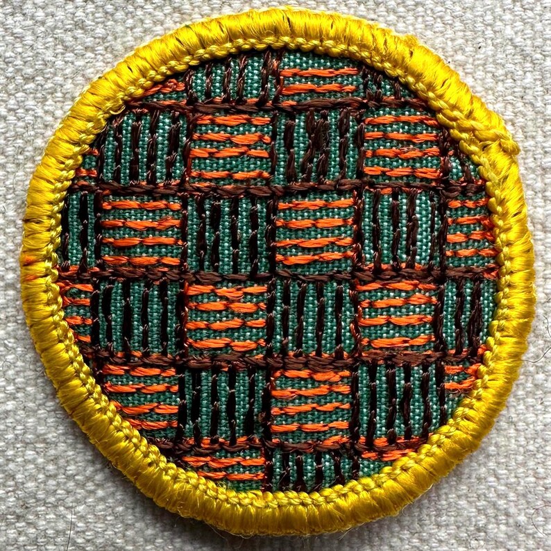 May include: A circular woven patch with a geometric design. The pattern includes a grid of teal and orange squares, framed by a brown weave. A yellow embroidered border surrounds the patch. Suitable for clothing or accessories.