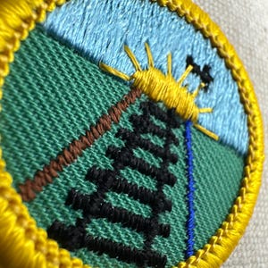 May include: Embroidered patch with a nature scene. The patch features a yellow sun, a black tree, and a brown diagonal line against a green background. A blue sky and a yellow border complete the design.