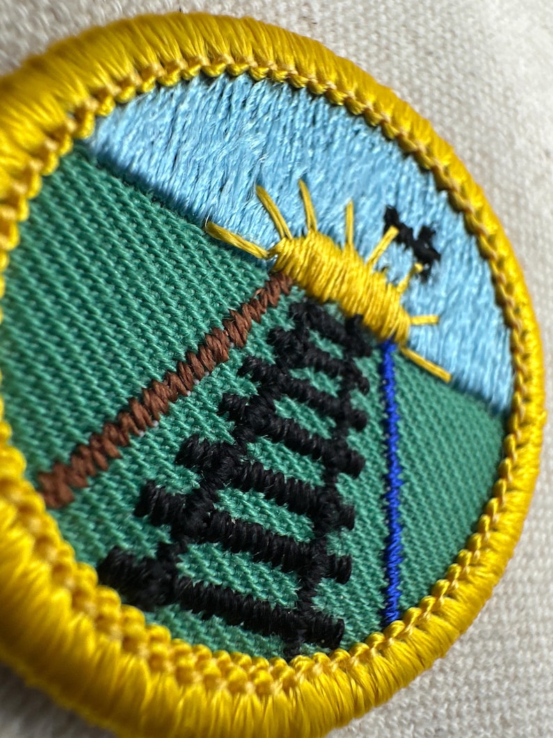 May include: Embroidered patch with a nature scene. The patch features a yellow sun, a black tree, and a brown diagonal line against a green background. A blue sky and a yellow border complete the design.