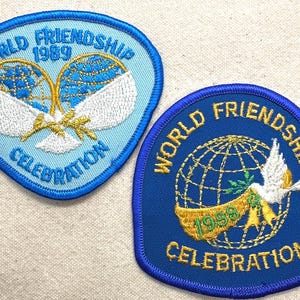 May include: Two embroidered patches with the words "World Friendship Celebration" and the year 1989 and 1988. The patches feature a globe, doves, and olive branches in blue, gold, and white.