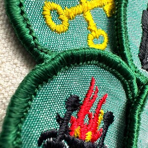 May include: Close-up of embroidered patches with green borders. One patch features a yellow key design, while another displays a black and red campfire. The patches are on a textured green fabric, showcasing detailed stitching.