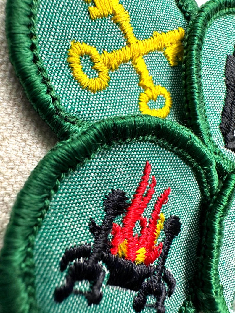 May include: Close-up of embroidered patches with green borders. One patch features a yellow key design, while another displays a black and red campfire. The patches are on a textured green fabric, showcasing detailed stitching.