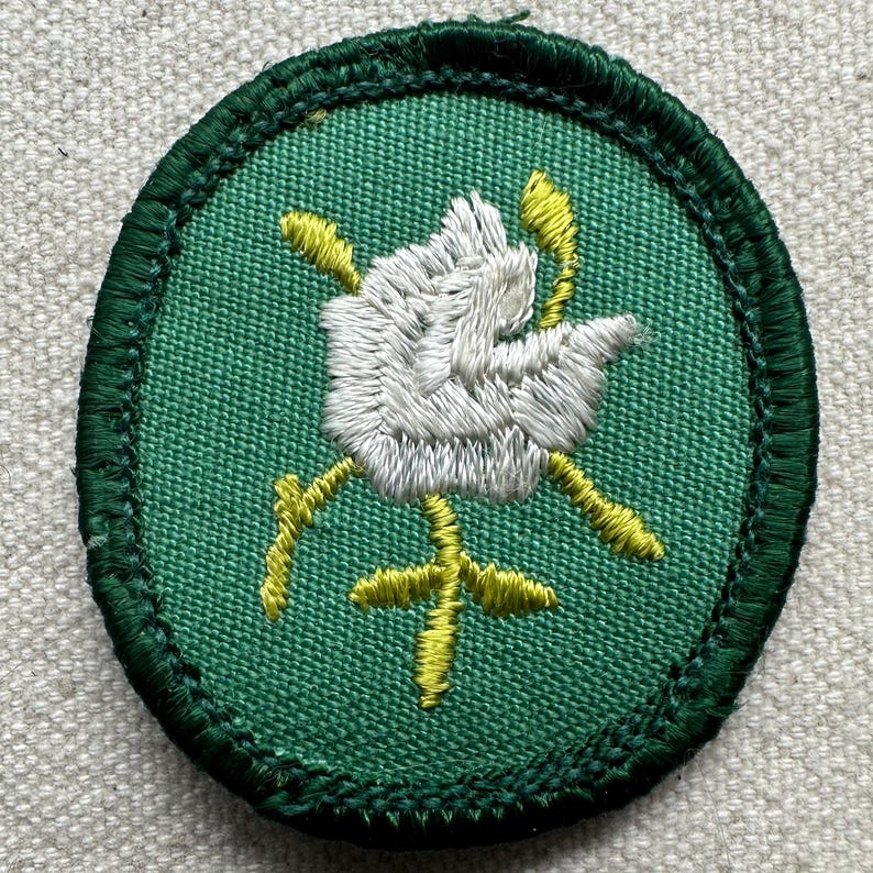 Pode incluir: Patch bordado com uma rosa branca e detalhes amarelos sobre fundo verde. O patch tem uma borda verde escura. O design provavelmente se destina a ser costurado em tecido. A rosa &eacute; o elemento central.