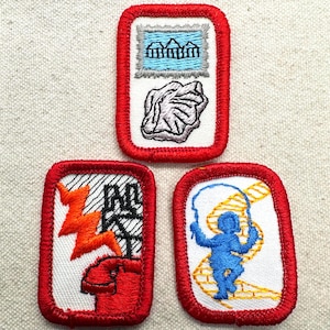May include: Three embroidered patches with red borders. One patch features a blue sky with three buildings and a gray rock. Another shows an orange lightning bolt and the letters "AD" and "KT". The third depicts a blue figure jumping rope.