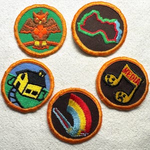 May include: A collection of embroidered patches with various designs. One patch features an owl, others depict a map, a house, a rainbow, and musical notes with the word "MEDIA". Each patch has a brown background and orange border.