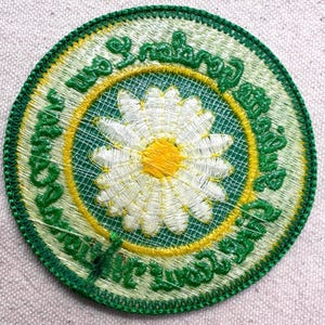 Puede incluir: Parche bordado redondo con una margarita blanca y centro amarillo. El dise&ntilde;o est&aacute; enmarcado por un c&iacute;rculo verde y amarillo, con texto verde alrededor del borde y un borde verde. El parche es redondo.