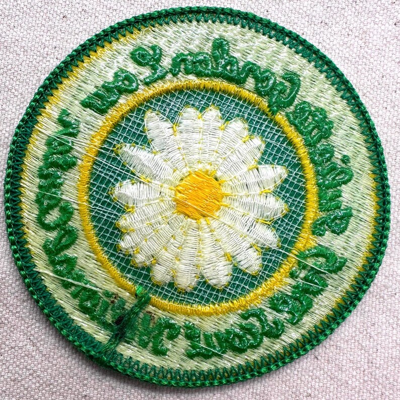 Puede incluir: Parche bordado redondo con una margarita blanca y centro amarillo. El dise&ntilde;o est&aacute; enmarcado por un c&iacute;rculo verde y amarillo, con texto verde alrededor del borde y un borde verde. El parche es redondo.