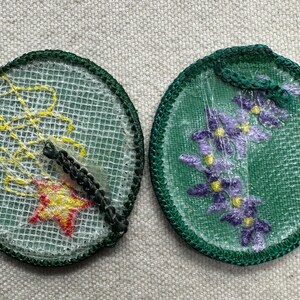 May include: Two round embroidered patches. One features a yellow and red star with yellow lines on a white mesh background, and a black border. The other has purple flowers on a green mesh background with a green border.
