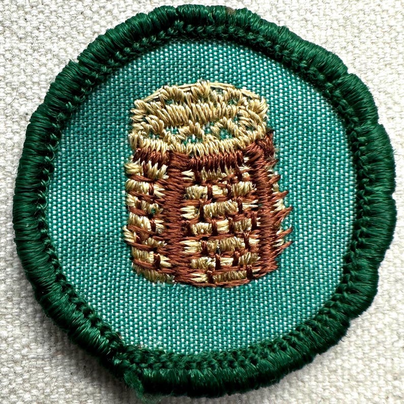 May include: A round, embroidered patch featuring a detailed owl design. The owl is rendered in shades of brown and tan, with a textured appearance. The patch has a teal background and a dark green, stitched border.