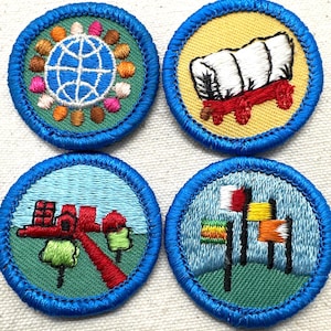 May include: Four embroidered patches with blue borders. Designs include a globe surrounded by colorful shapes, a covered wagon, a red structure with trees, and flags. These patches are likely for a youth organization.