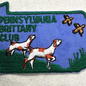 May include: Embroidered patch in the shape of Pennsylvania, featuring the text "PENNSYLVANIA BRITTANY CLUB". The patch depicts two Brittany Spaniels and two birds in flight, with green and orange accents on a blue background.