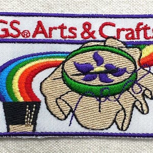 Puede incluir: Parche bordado con el texto "GS Arts & Crafts" en rojo. El parche presenta un arco&iacute;ris, una mano que sostiene un dise&ntilde;o verde y morado, y un borde morado.