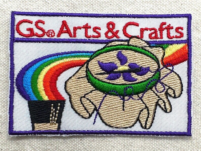 Puede incluir: Parche bordado con el texto "GS Arts & Crafts" en rojo. El parche presenta un arco&iacute;ris, una mano que sostiene un dise&ntilde;o verde y morado, y un borde morado.