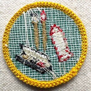 May include: Embroidered patch with a yellow border, featuring a feather, two torches, and a wrapped item. The background is green with a white grid pattern. The feather has black, white, and red details. The torches are tan, and the wrapped item is white with red accents.