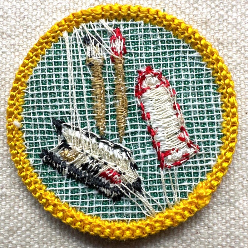 May include: Embroidered patch with a yellow border, featuring a feather, two torches, and a wrapped item. The background is green with a white grid pattern. The feather has black, white, and red details. The torches are tan, and the wrapped item is white with red accents.