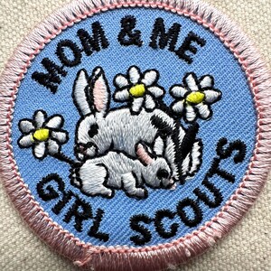 May include: Embroidered patch with the text "MOM & ME GIRL SCOUTS". The patch features two rabbits, one larger and one smaller, with white fur and black markings, surrounded by daisies with yellow centers. The patch has a light blue background with a pink border.
