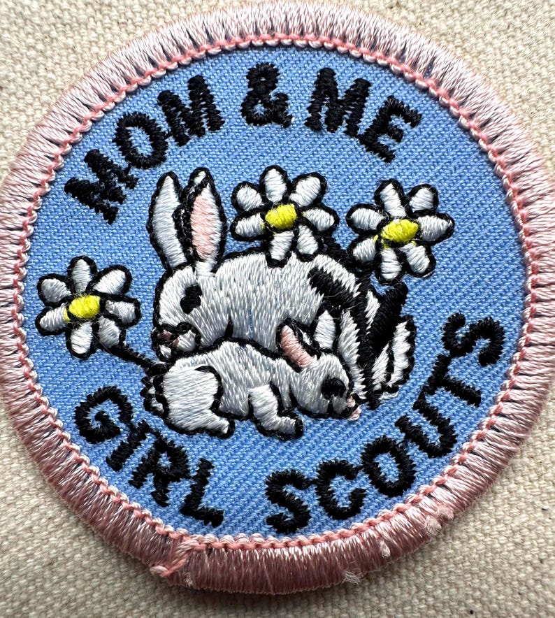May include: Embroidered patch with the text "MOM & ME GIRL SCOUTS". The patch features two rabbits, one larger and one smaller, with white fur and black markings, surrounded by daisies with yellow centers. The patch has a light blue background with a pink border.