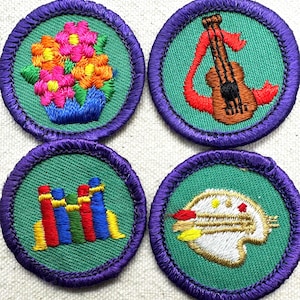 May include: Four embroidered patches with a green background and purple trim. The patches feature a bouquet of flowers, a guitar, colorful shapes, and a bird with a paintbrush. The patches are round.