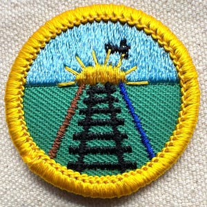 May include: Embroidered patch with a yellow border. The patch features a blue sky, a green field, a black tower, and a yellow sun with rays. Brown and blue lines extend from the tower. The patch is round.