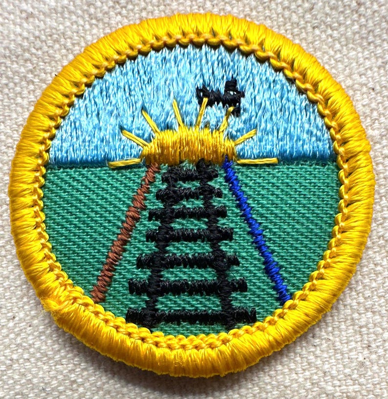 May include: Embroidered patch with a yellow border. The patch features a blue sky, a green field, a black tower, and a yellow sun with rays. Brown and blue lines extend from the tower. The patch is round.