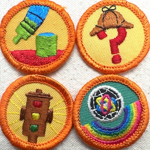 May include: Four round embroidered patches with orange borders. The patches feature various designs: a paintbrush and paint can, a detective hat and question mark, a traffic light, and a rainbow with a central emblem. The backgrounds are yellow and green.