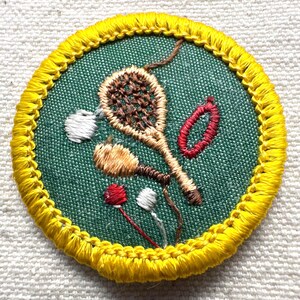 May include: Embroidered patch with a green background and a yellow border. The patch features brown, red, and white embroidered designs of a tennis racket, a shuttlecock, and other sports-related elements. The patch is round.