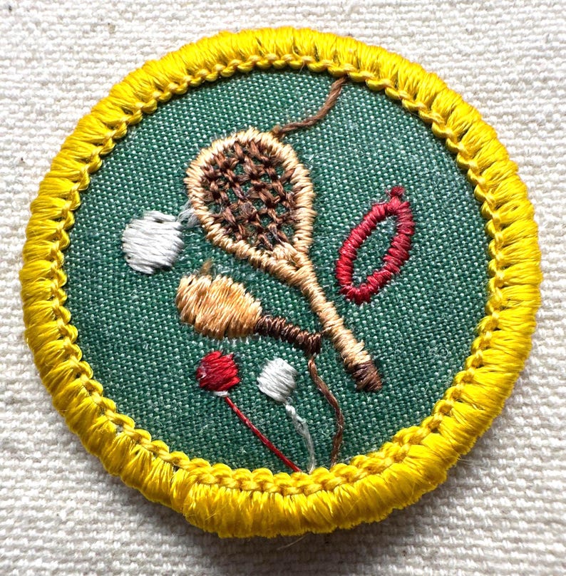 May include: Embroidered patch with a green background and a yellow border. The patch features brown, red, and white embroidered designs of a tennis racket, a shuttlecock, and other sports-related elements. The patch is round.