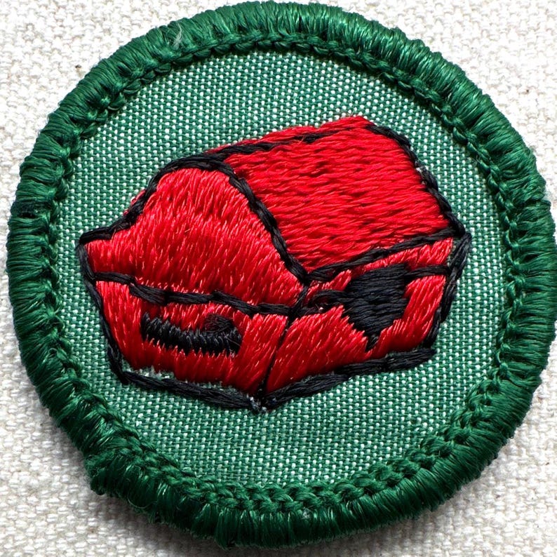 May include: Embroidered patch featuring a red object with black accents, possibly a building or container, on a green background. The patch has a green border and is likely for a uniform or craft project.